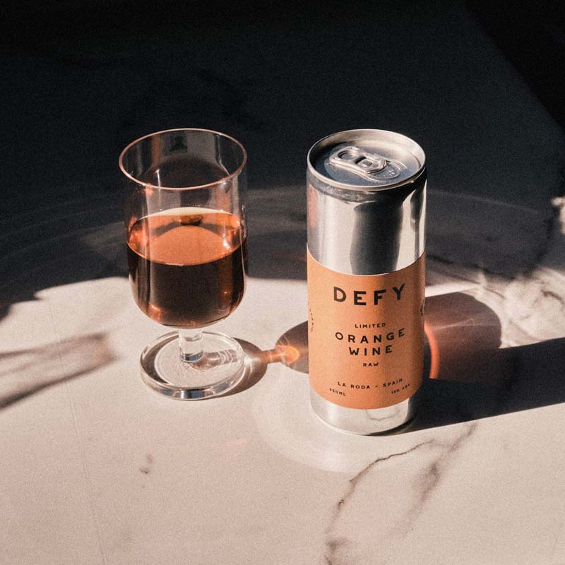 A can of DEFY premium Spanish vegan organic orange wine next to a glass of the orange wine, in the sunshine.
